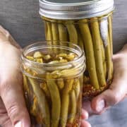 Mike from Chili Pepper Madness holds two jars of Pickled Green Beans (Spicy Dilly Beans) in brine, with vibrant spices visible at the bottom.