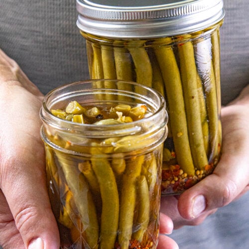 Mike from Chili Pepper Madness holds two jars of Pickled Green Beans (Spicy Dilly Beans) in brine, with vibrant spices visible at the bottom.