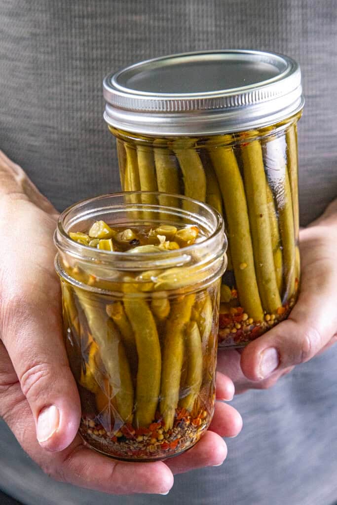 Mike from Chili Pepper Madness holds two jars of Pickled Green Beans (Spicy Dilly Beans) in brine, with vibrant spices visible at the bottom.