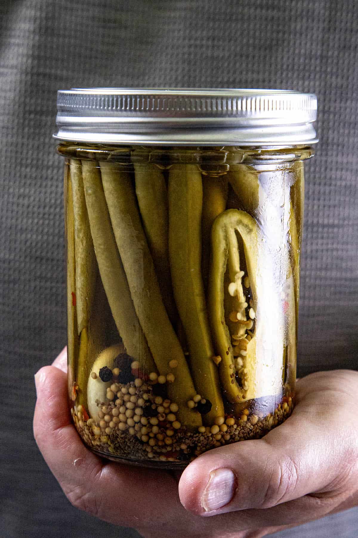 A hand holds a glass jar filled with Pickled Green Beans (Spicy Dilly Beans), brimming with spices and seeds, and sealed tightly with a metal lid.