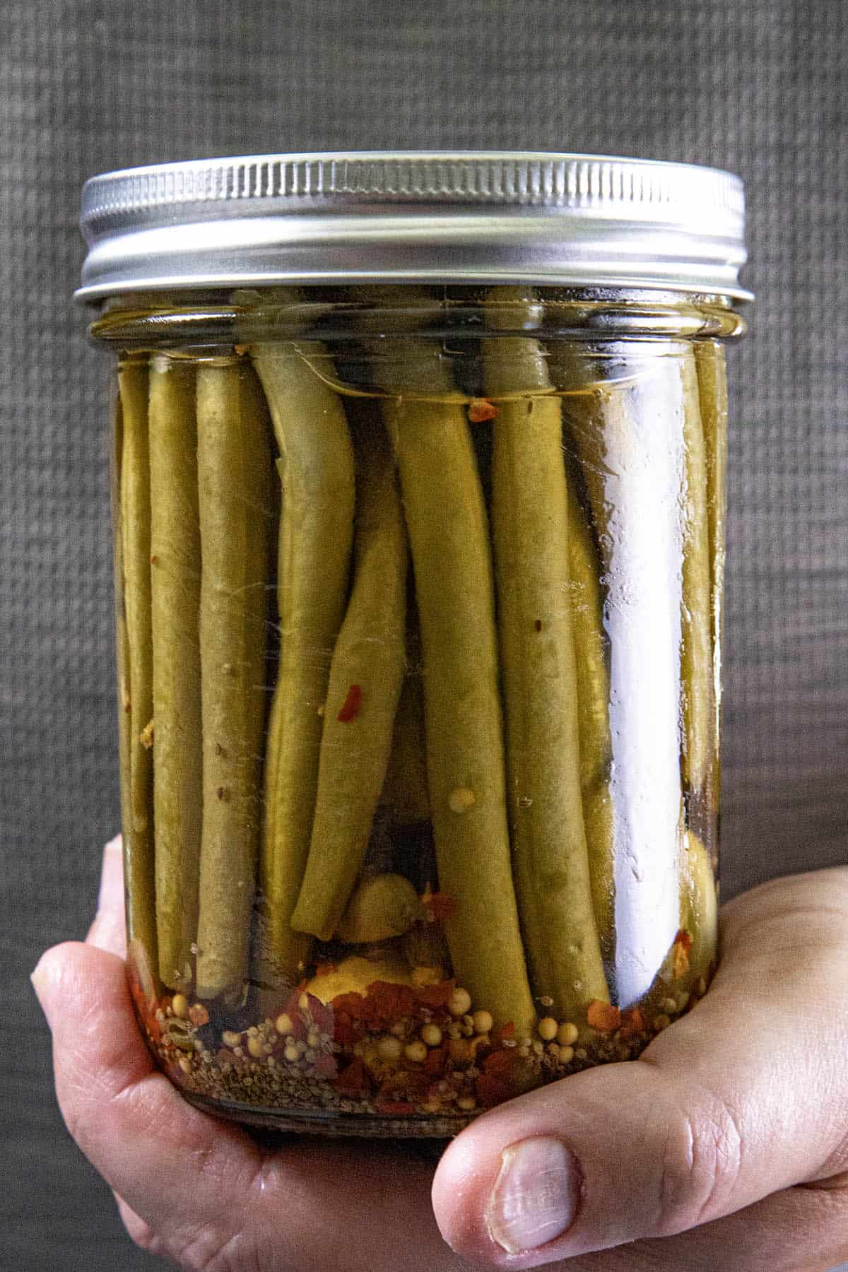 A hand holds a glass jar filled with Pickled Green Beans, spices, and seeds in brine, against a textured gray background.