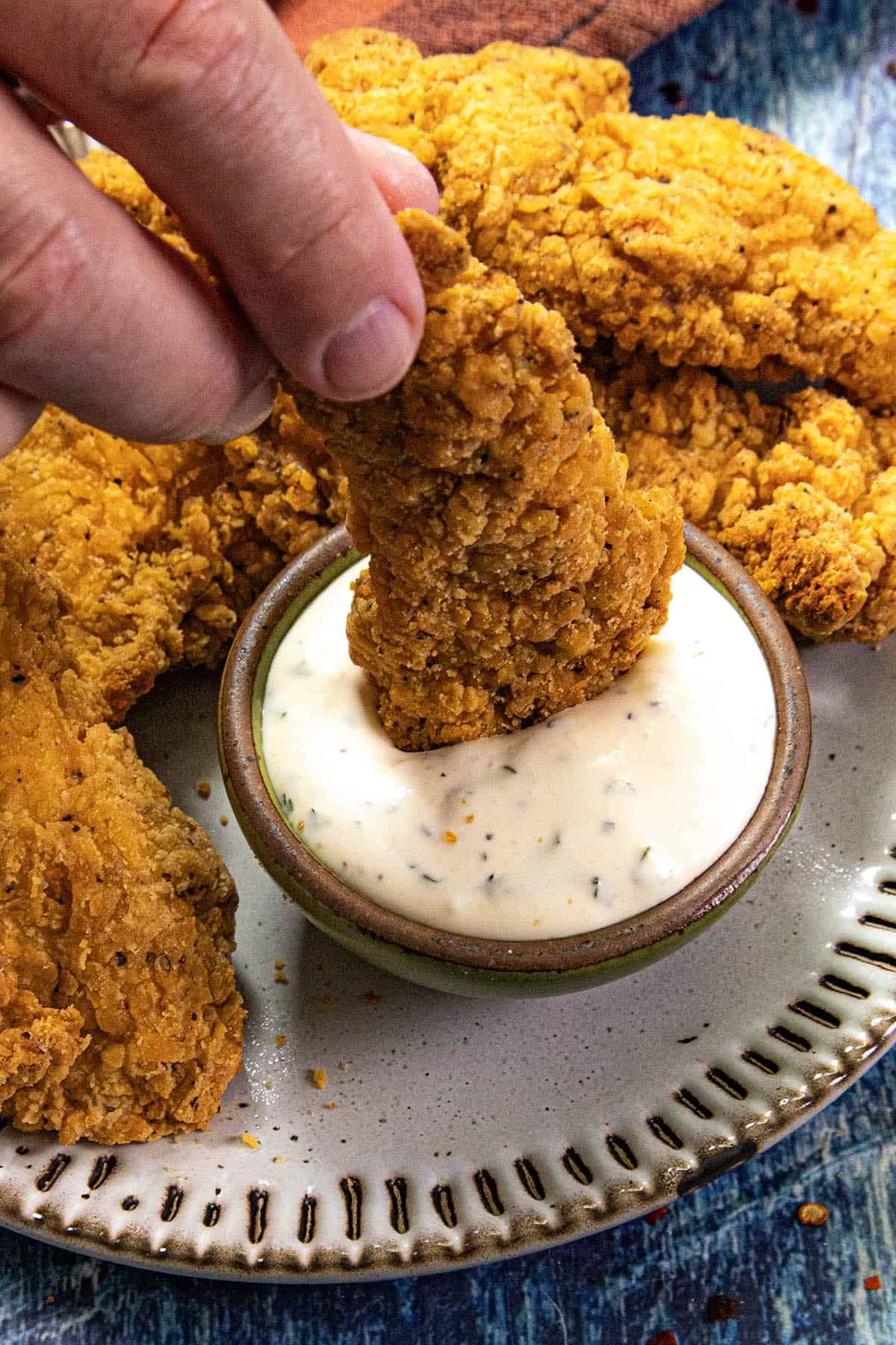 A hand dips a crispy fried chicken tender into a small bowl of creamy homemade ranch dressing, with more chicken tenders on a plate in the background.