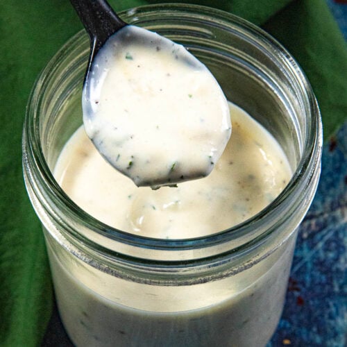 A spoon holds up creamy Homemade Ranch Dressing over a glass jar filled with more dressing, set on a blue surface with a green cloth nearby.