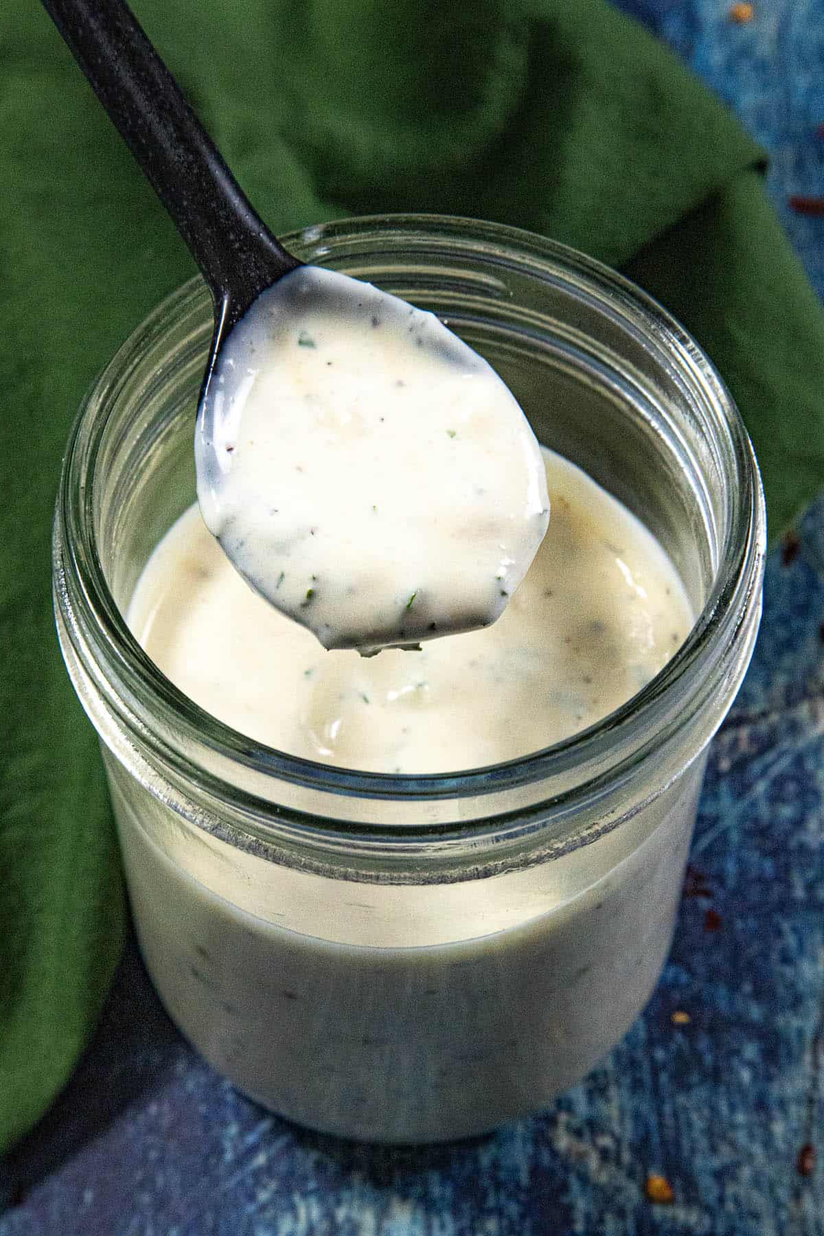 A spoon holds up creamy Homemade Ranch Dressing over a glass jar filled with more dressing, set on a blue surface with a green cloth nearby.