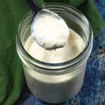 A spoon holds up creamy homemade ranch dressing above an open glass jar filled with more dressing, set on a dark blue surface with a green cloth nearby.