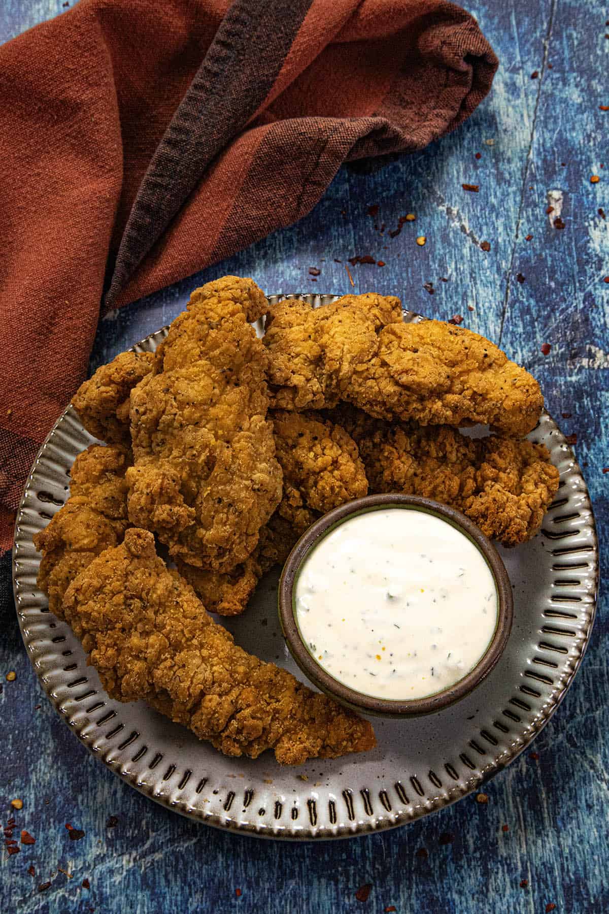 A plate of crispy fried chicken tenders with a cup of creamy homemade ranch dressing sits on a blue textured surface, next to a folded rust-colored cloth napkin.