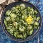 A dark plate of thinly sliced Japanese Cucumber Salad (Sunomono), topped with sesame seeds, red pepper flakes, and a small mound of grated ginger, set on a blue wooden table.