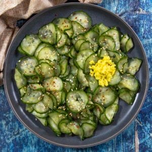 A dark plate of thinly sliced Japanese Cucumber Salad (Sunomono), topped with sesame seeds, red pepper flakes, and a small mound of grated ginger, set on a blue wooden table.