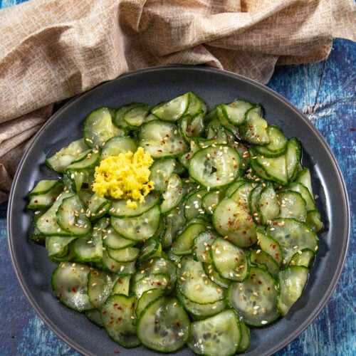 A black plate of thinly sliced Japanese Cucumber Salad (Sunomono) with sesame seeds, red pepper flakes, and a mound of grated ginger, on a blue textured surface beside a beige cloth.