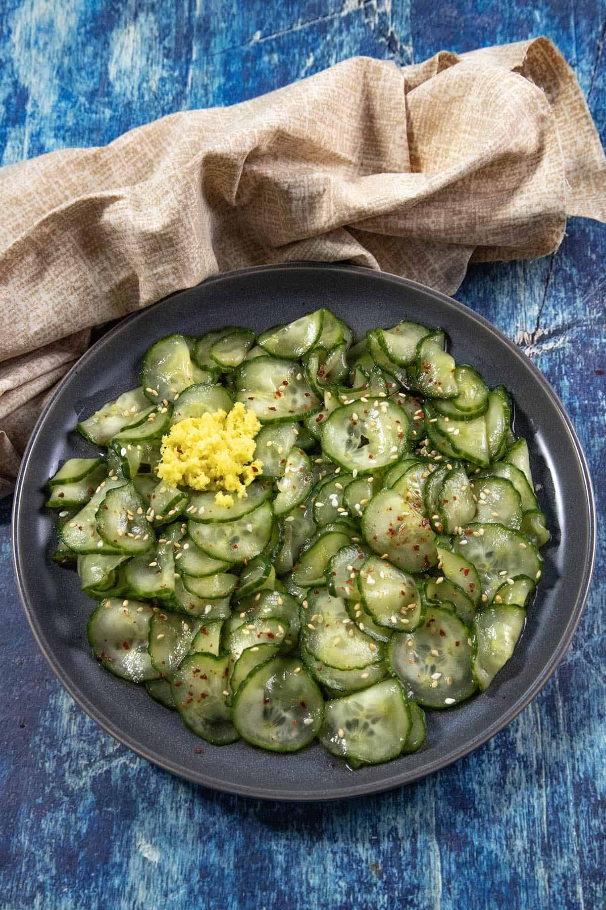 A black plate of thinly sliced Japanese Cucumber Salad (Sunomono) with sesame seeds, red pepper flakes, and a mound of grated ginger, on a blue textured surface beside a beige cloth.