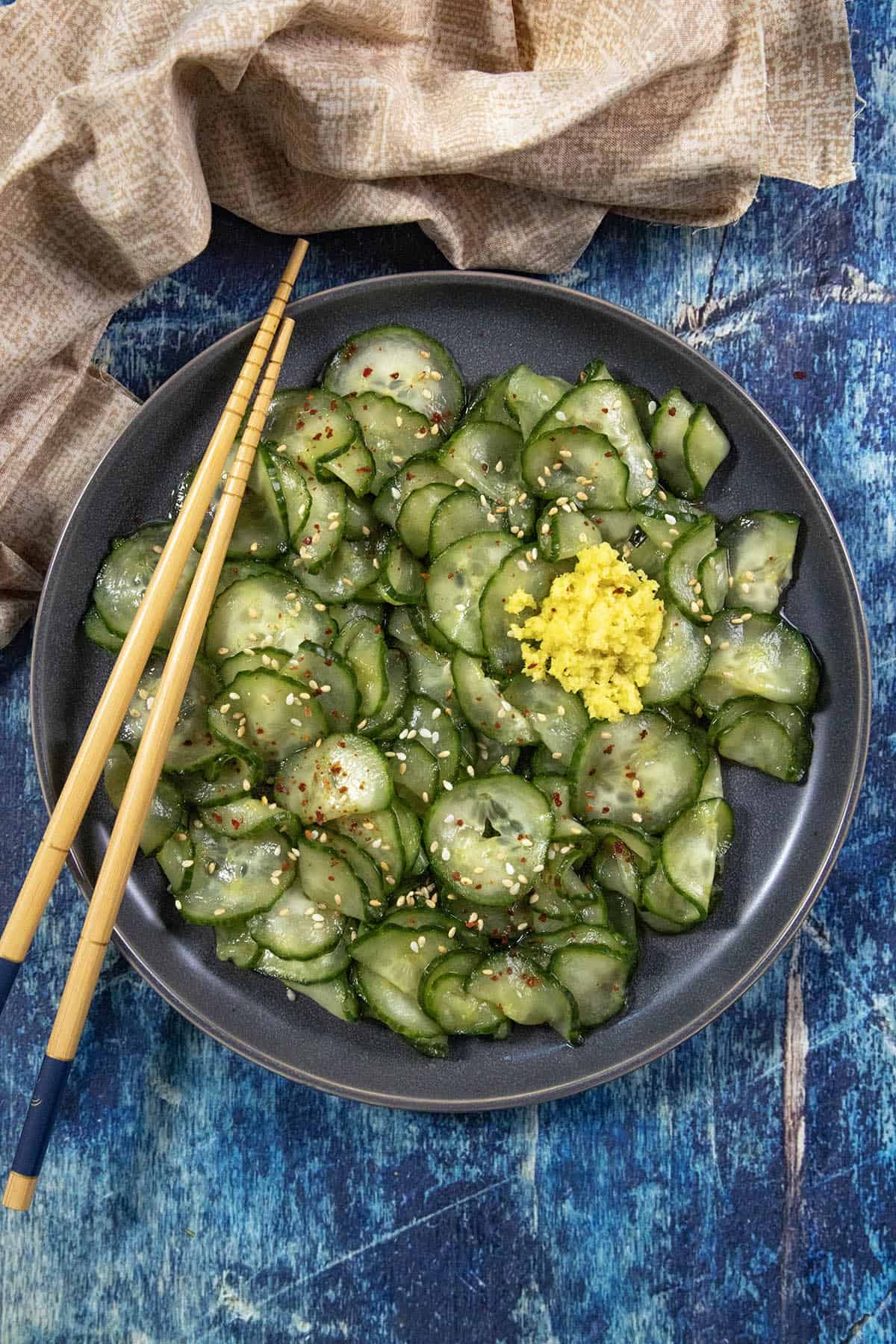 A black plate of Japanese Cucumber Salad (Sunomono), featuring sliced cucumber garnished with sesame seeds and a mound of grated ginger, sits invitingly with wooden chopsticks resting on the side, all set atop a blue rustic table.