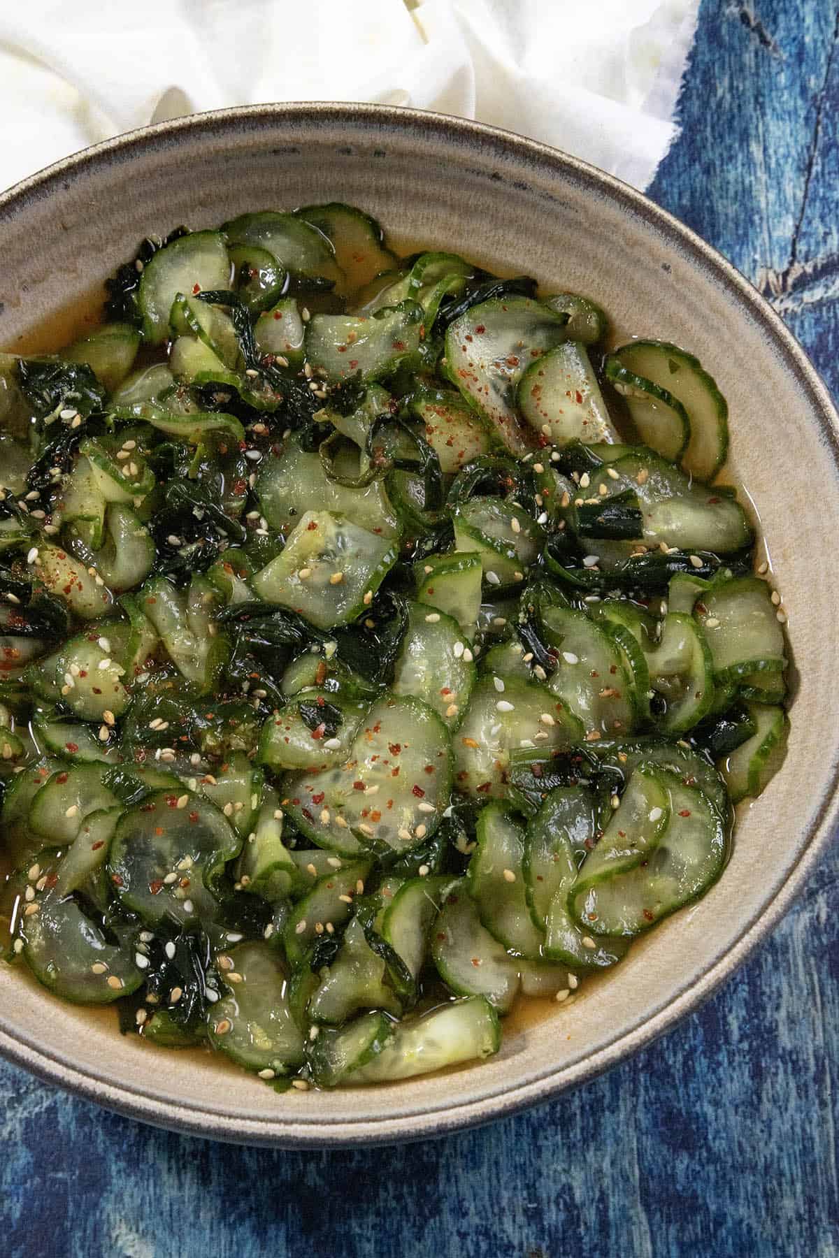 A bowl of sunomono (Japanese cucumber salad) with sliced cucumbers and seaweed in a light dressing, sprinkled with sesame seeds and red pepper flakes, rests on a blue textured surface.
