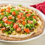 Taco dip spread along a serving dish with layered refried beans, cream cheese, diced tomatoes and more