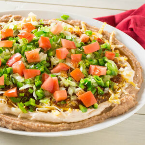 Taco dip spread along a serving dish with layered refried beans, cream cheese, diced tomatoes and more