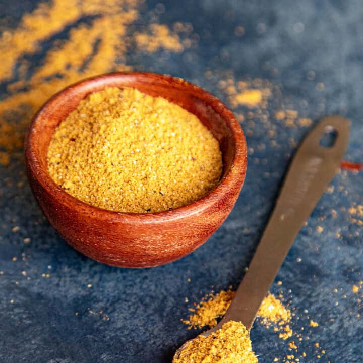 A small wooden bowl filled with homemade Vindaloo Curry Powder. Beside it, a metal measuring spoon holds a scoop of the same vindaloo masala, with some spilled on the surface around both.
