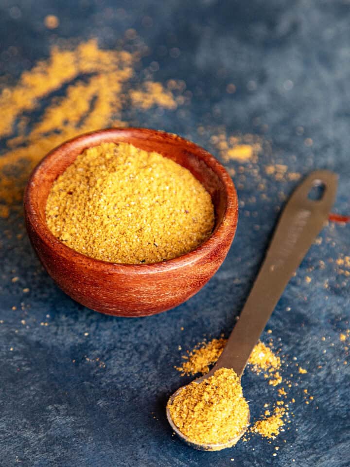 A small wooden bowl filled with homemade Vindaloo Curry Powder. Beside it, a metal measuring spoon holds a scoop of the same vindaloo masala, with some spilled on the surface around both.