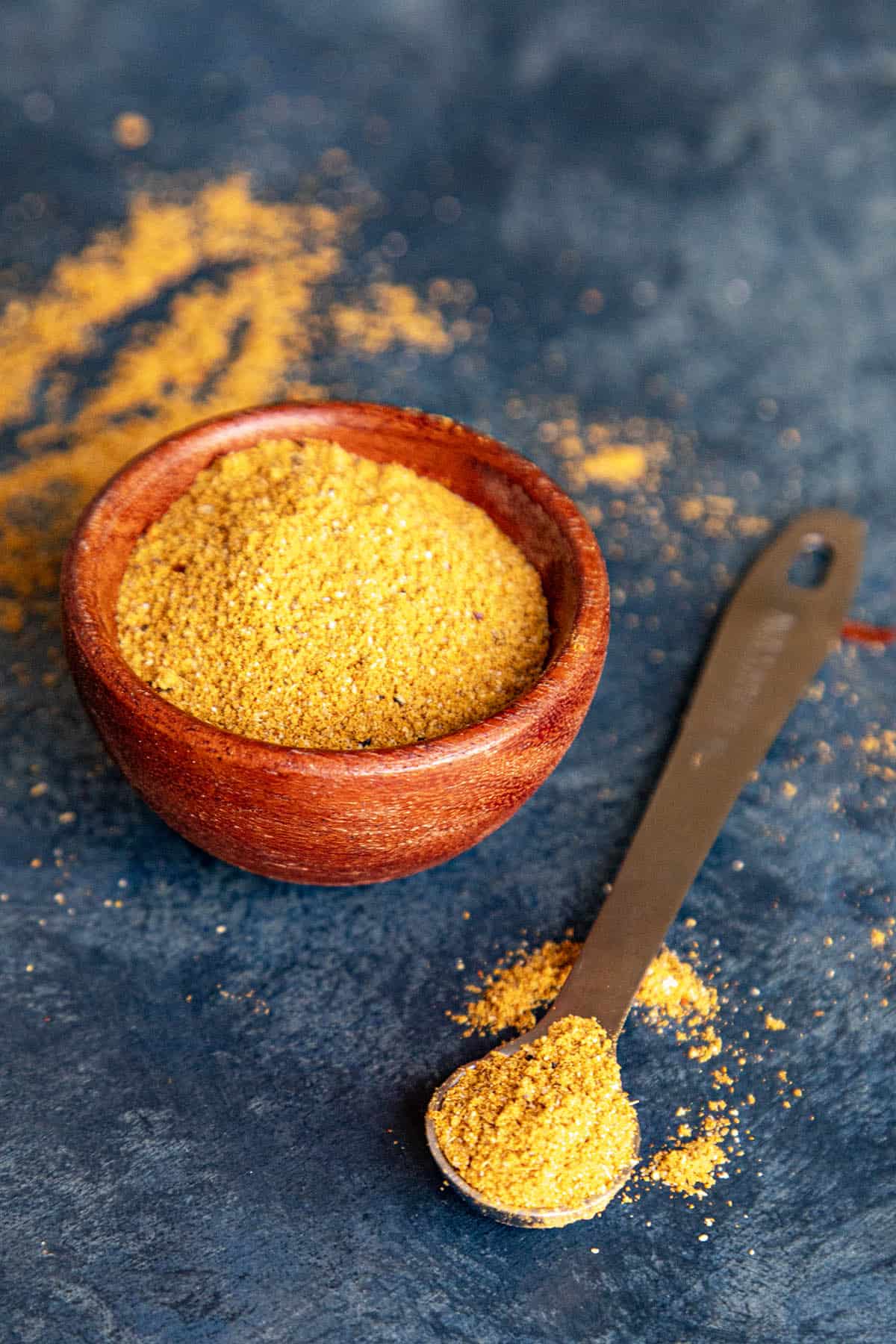 A small wooden bowl filled with homemade Vindaloo Curry Powder. Beside it, a metal measuring spoon holds a scoop of the same vindaloo masala, with some spilled on the surface around both.
