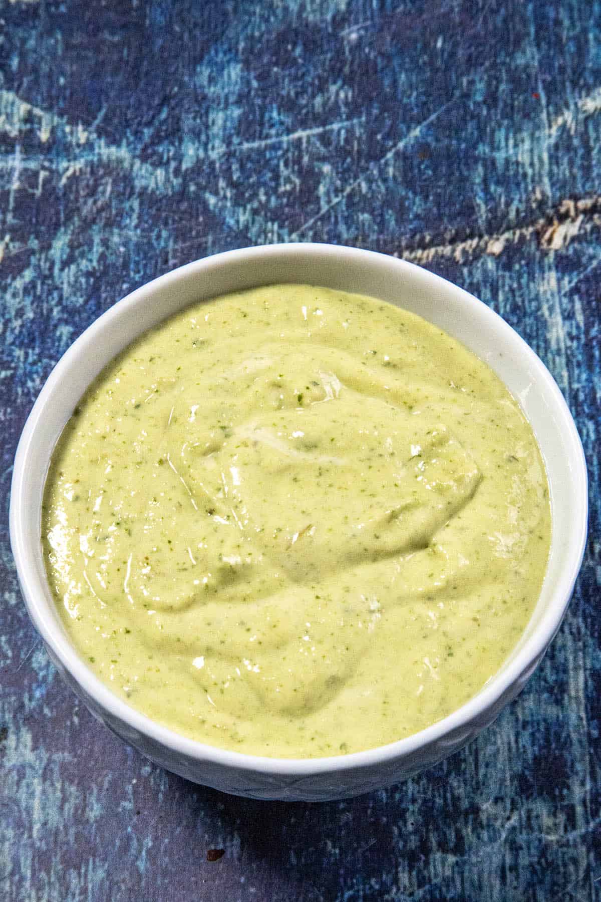 A white bowl filled with creamy green sauce, perfect as an easy Jalapeno Crema for breakfast burritos, sits on a textured blue surface.
