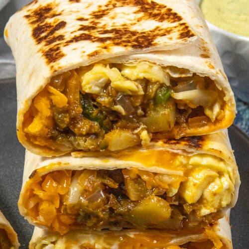 Three Easy Breakfast Burritos are stacked on a plate, each cut in half to reveal a filling of scrambled eggs, potatoes, cheese, and vegetables. A bowl of green sauce is in the background.
