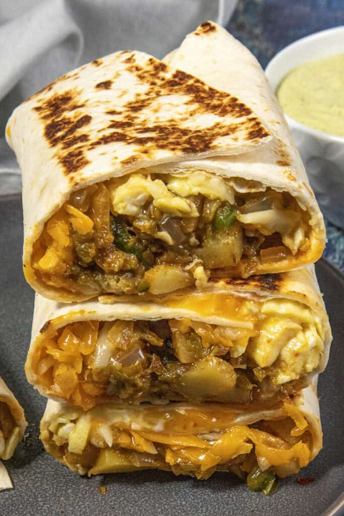 Three Easy Breakfast Burritos are stacked on a plate, each cut in half to reveal a filling of scrambled eggs, potatoes, cheese, and vegetables. A bowl of green sauce is in the background.