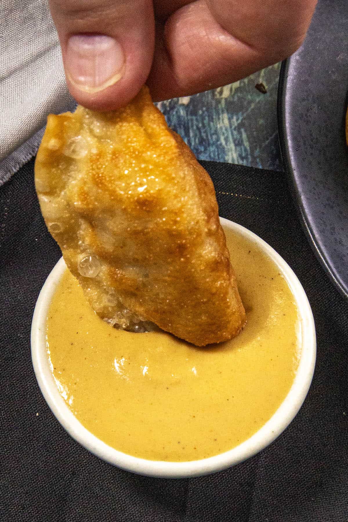 A hand dips a fried dumpling into a small bowl of creamy, yellow Chinese Hot Mustard dipping sauce.
