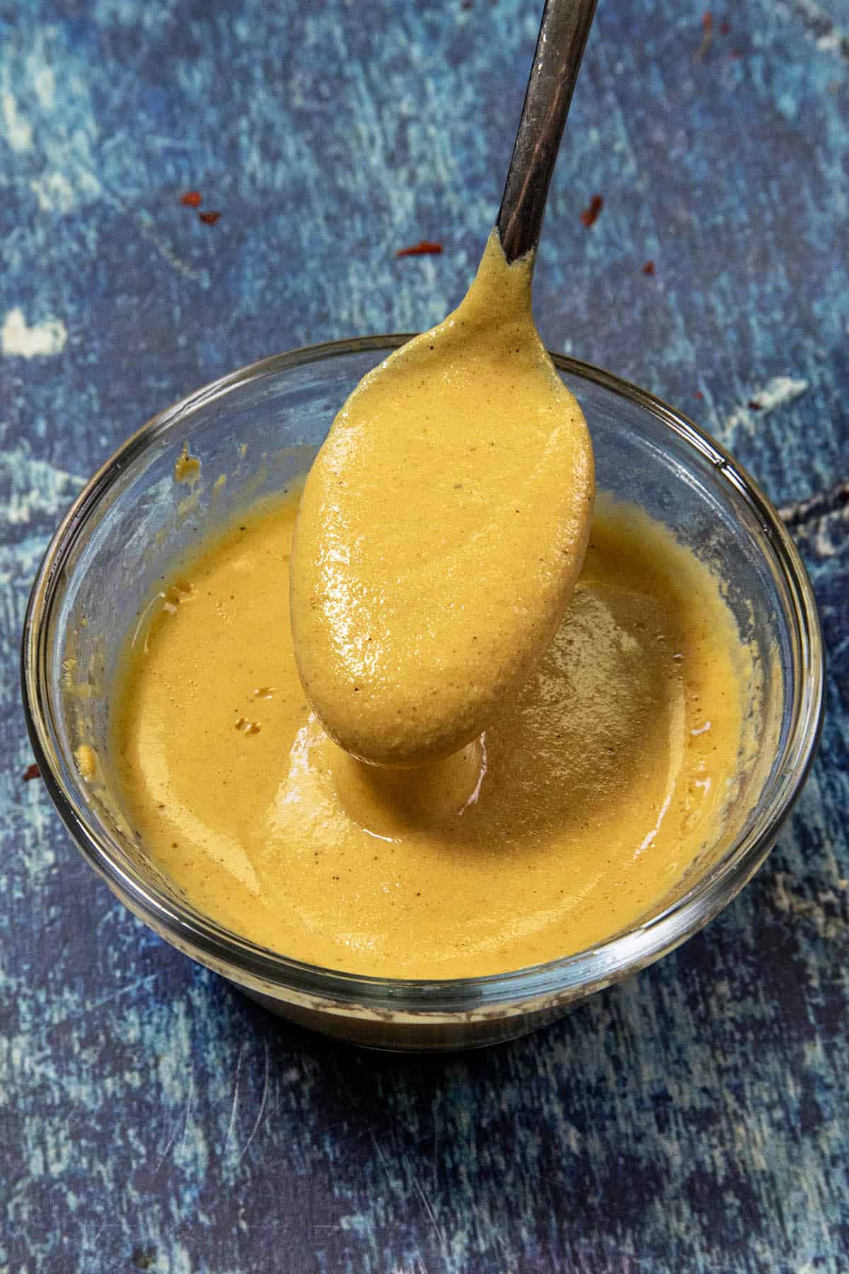 A glass bowl filled with thick yellow Chinese Hot Mustard sauce sits in focus. A spoon dipped in the sauce is being held above the bowl, with the tangy condiment coating the spoon. The background features a blue, textured surface.