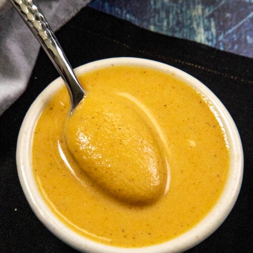 A small white bowl filled with creamy, golden-brown Chinese hot mustard sauce, with a metal spoon resting inside. The bowl is placed on a dark surface with a white cloth nearby.