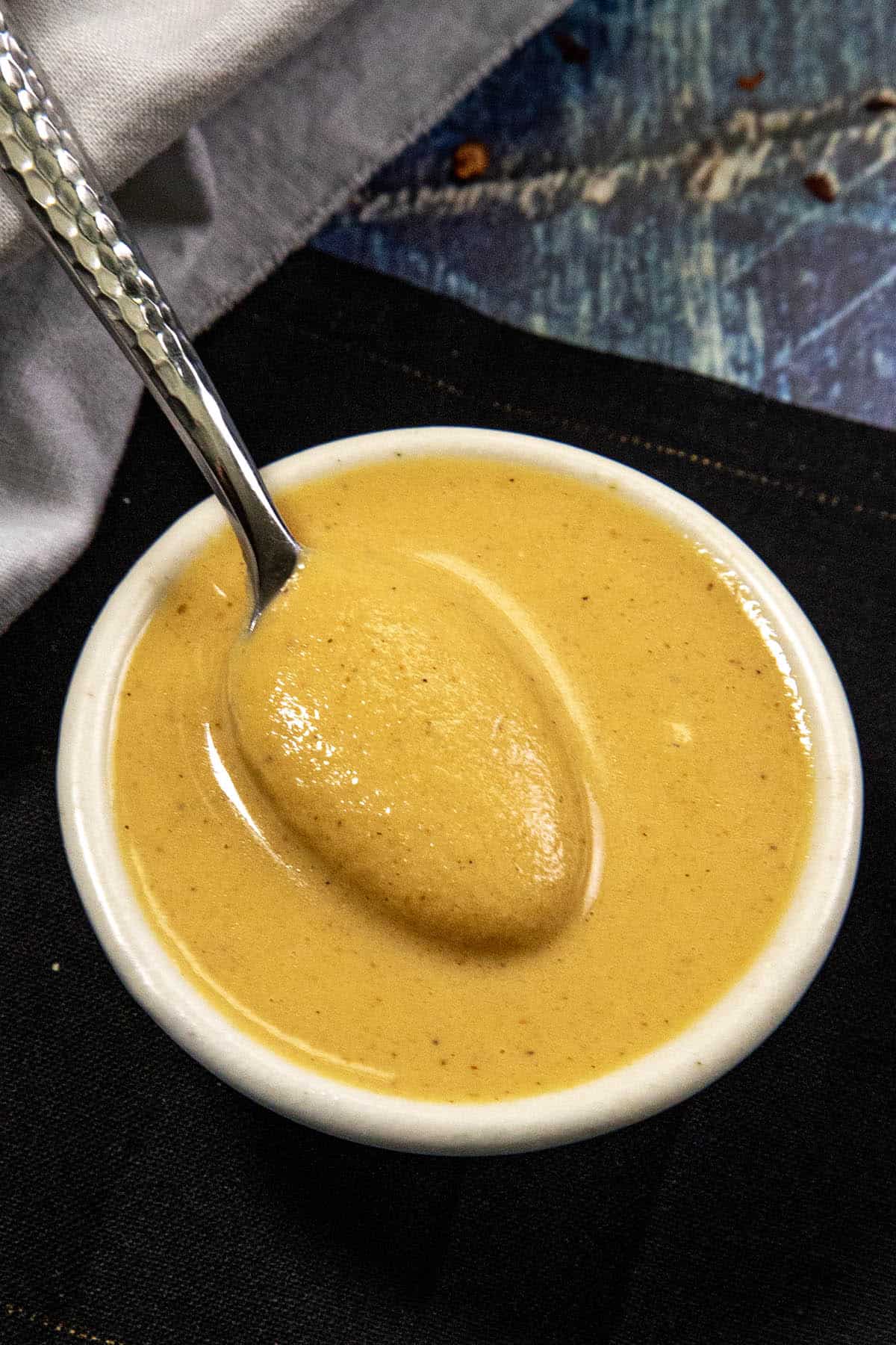 A small white bowl filled with creamy, golden-brown Chinese hot mustard sauce, with a metal spoon resting inside. The bowl is placed on a dark surface with a white cloth nearby.