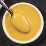 A small white bowl filled with creamy yellow Chinese Hot Mustard sauce, with a spoon resting inside, on a dark fabric background.