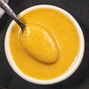 A small white bowl filled with creamy yellow Chinese Hot Mustard sauce, with a spoon resting inside, on a dark fabric background.