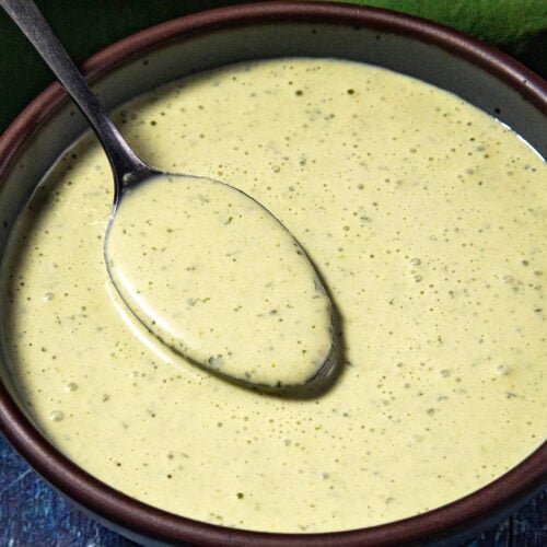 A bowl of creamy green Jalapeno Crema with herbs, shown with a spoon resting inside. The background includes a green cloth on a blue textured surface.