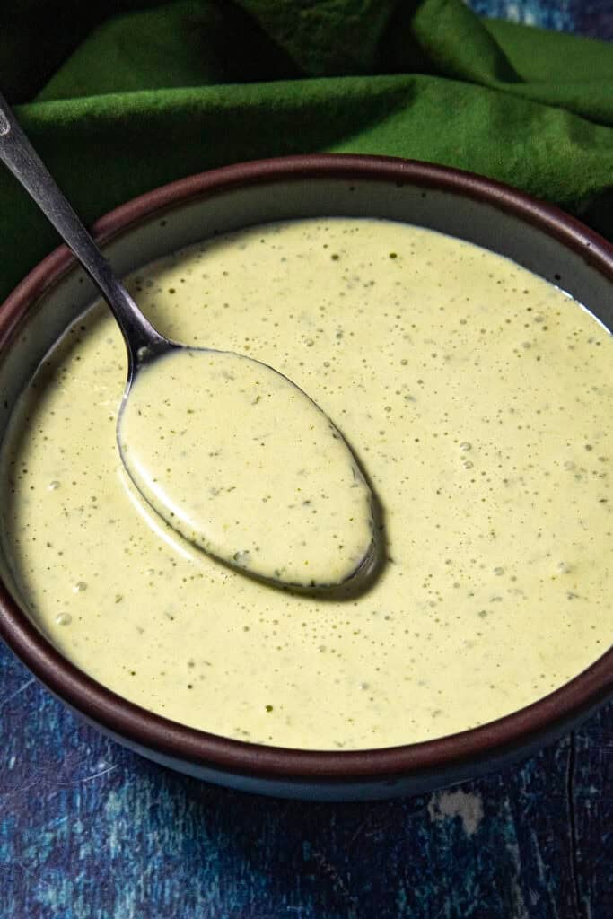 A bowl of creamy green Jalapeno Crema with herbs, shown with a spoon resting inside. The background includes a green cloth on a blue textured surface.