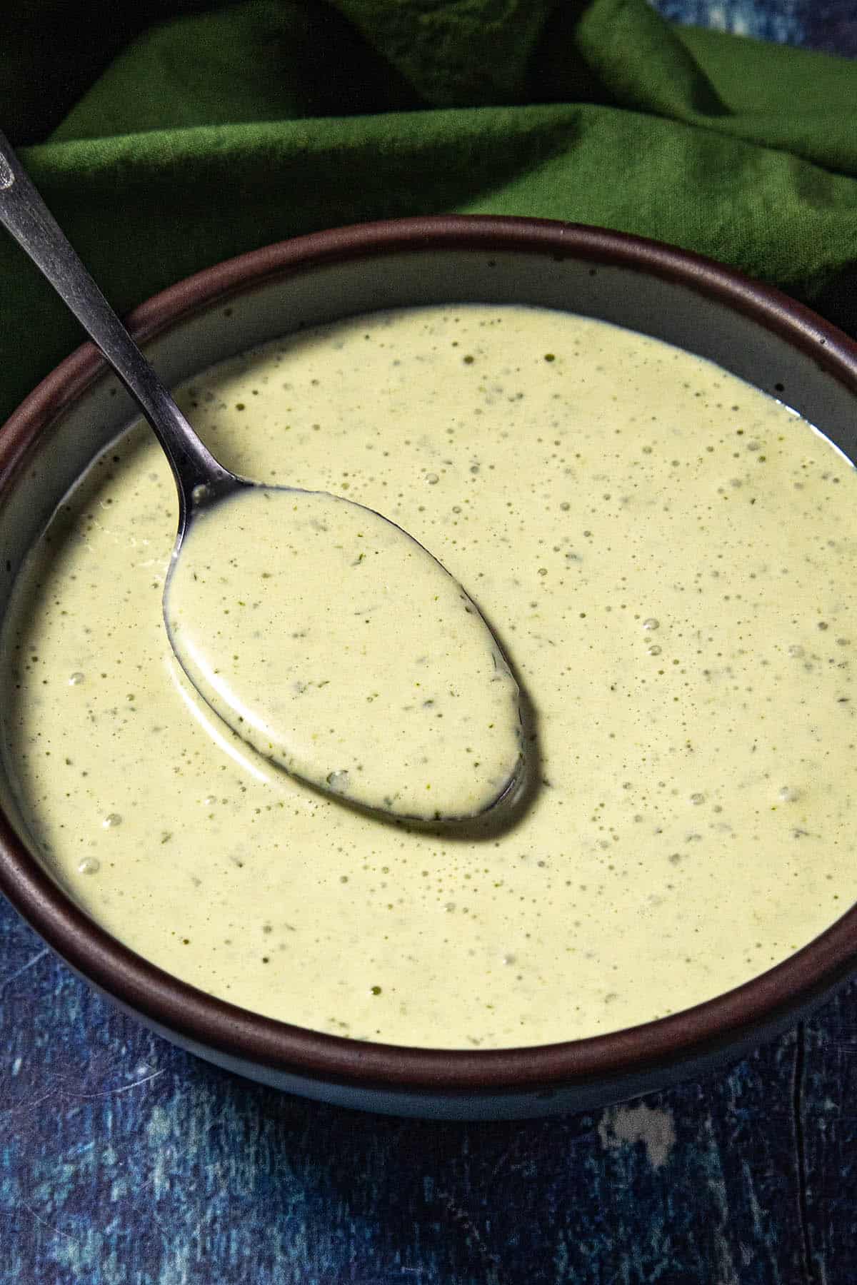 A bowl of creamy green Jalapeno Crema with herbs, shown with a spoon resting inside. The background includes a green cloth on a blue textured surface.