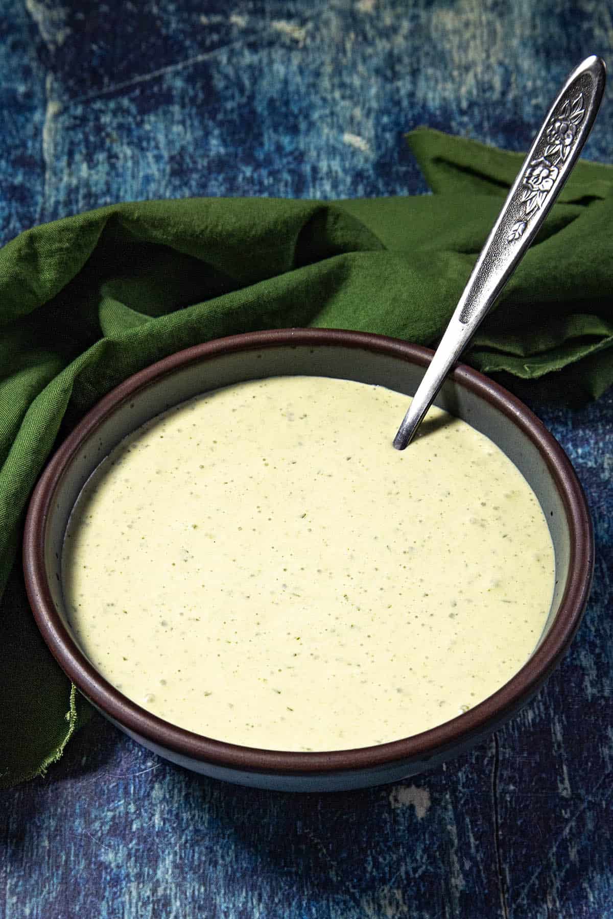 A bowl of creamy Spicy Jalapeno Crema with a spoon in it sits on a blue textured surface, next to a green cloth napkin.