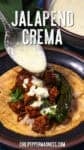 A spoon drizzles Spicy Jalapeno Crema over a taco filled with seasoned meat, onions, cilantro, and a whole roasted jalapeño, on a corn tortilla. Text reads Jalapeno Crema and chilipeppermadness.com.