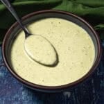 A spoon rests in a bowl filled with creamy green sauce (jalapeno crema) on a dark blue textured surface with a green cloth nearby.