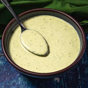 A spoon rests in a bowl filled with creamy green sauce (jalapeno crema) on a dark blue textured surface with a green cloth nearby.