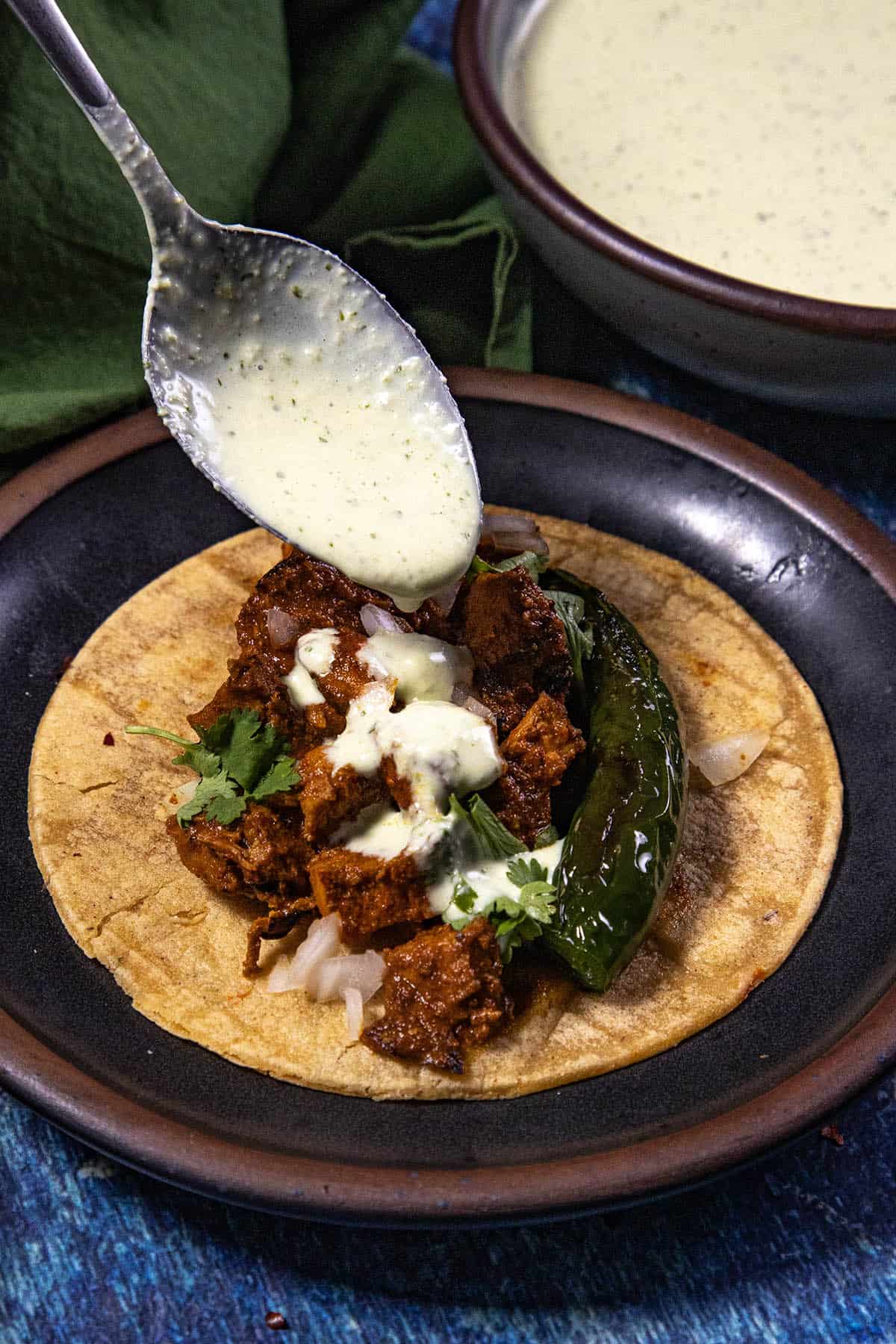 A spoon drizzles creamy Spicy Jalapeno Crema over a taco filled with marinated meat, onions, cilantro, and a whole grilled pepper, all on a corn tortilla on a rustic plate.