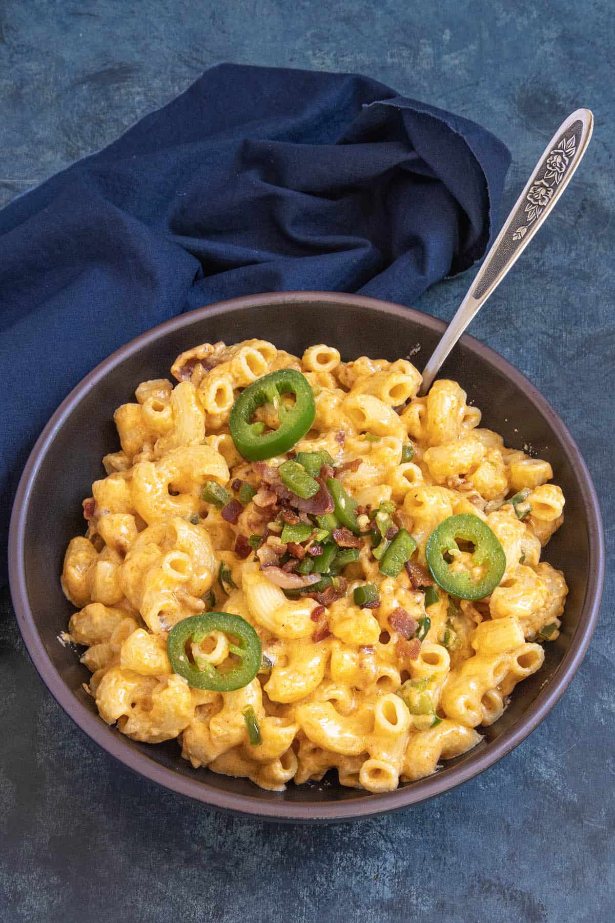 Creamy Jalapeno Popper Mac and Cheese - Recipe A bowl of creamy Jalapeno Popper Mac and Cheese topped with sliced jalapeños, chopped green onions, and bits of bacon, with a spoon and a dark blue napkin beside it.