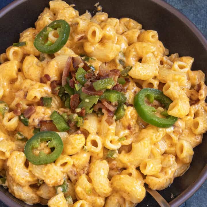 Indulge in a bowl of Jalapeno Popper Mac and Cheese, featuring creamy macaroni and cheese generously topped with sliced jalapeños, chopped bacon, and fresh green onions. A spoon rests inside the bowl, while a dark cloth is partially visible in the background.