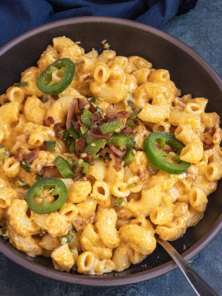 Indulge in a bowl of Jalapeno Popper Mac and Cheese, featuring creamy macaroni and cheese generously topped with sliced jalapeños, chopped bacon, and fresh green onions. A spoon rests inside the bowl, while a dark cloth is partially visible in the background.