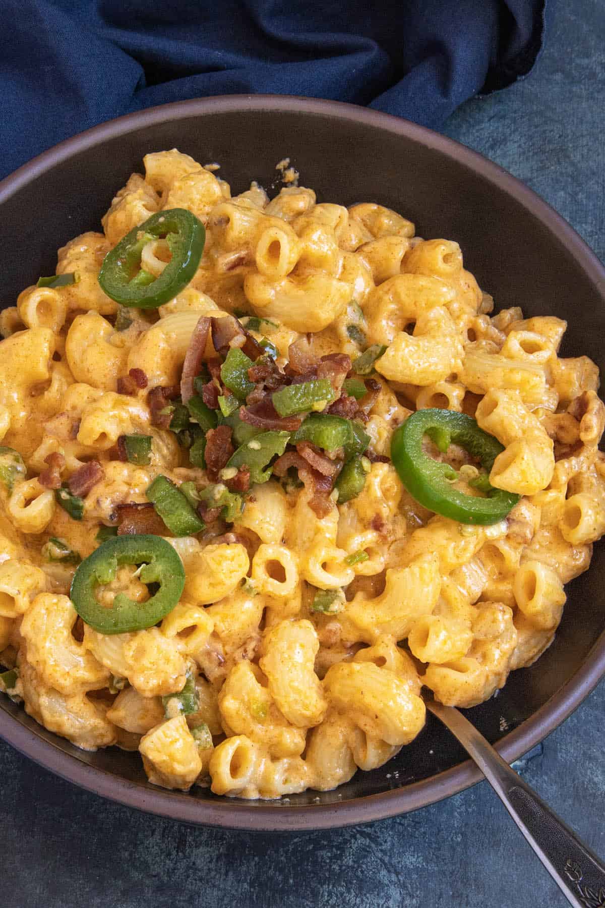 Indulge in a bowl of Jalapeno Popper Mac and Cheese, featuring creamy macaroni and cheese generously topped with sliced jalapeños, chopped bacon, and fresh green onions. A spoon rests inside the bowl, while a dark cloth is partially visible in the background.