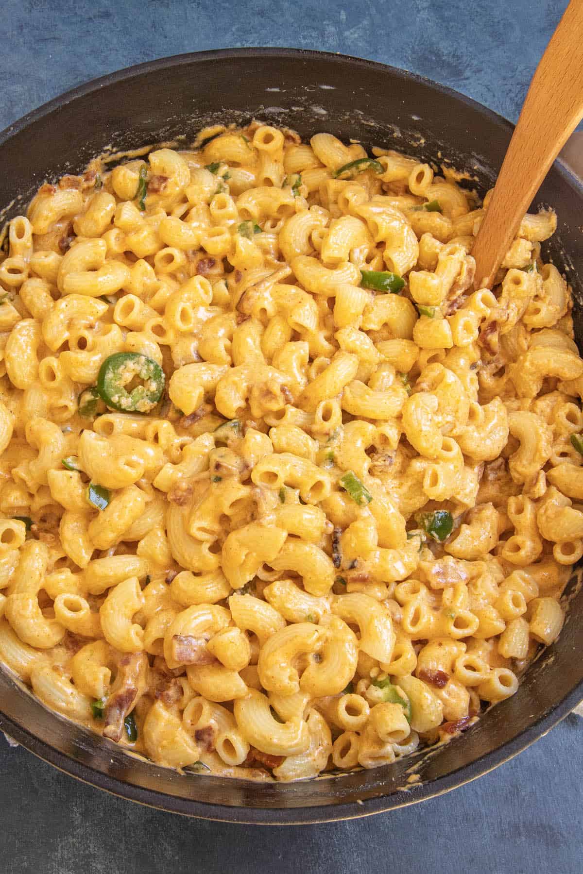 Creamy Jalapeno Popper Mac and Cheese with jalapeño slices and bits of bacon in a black skillet, stirred with a wooden spoon on a dark surface.