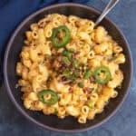A bowl of creamy Jalapeno Popper Mac and Cheese topped with sliced jalapeños and bits of crispy bacon, with a spoon on the side.
