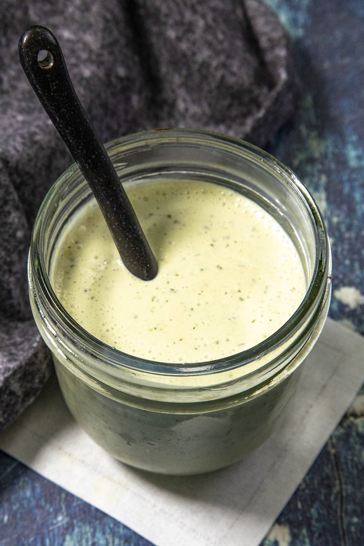 A glass jar filled with creamy green Jalapeno Ranch Dressing, with a black spoon inside, sits on a white square mat. A gray cloth is partially visible in the background on a blue textured surface.