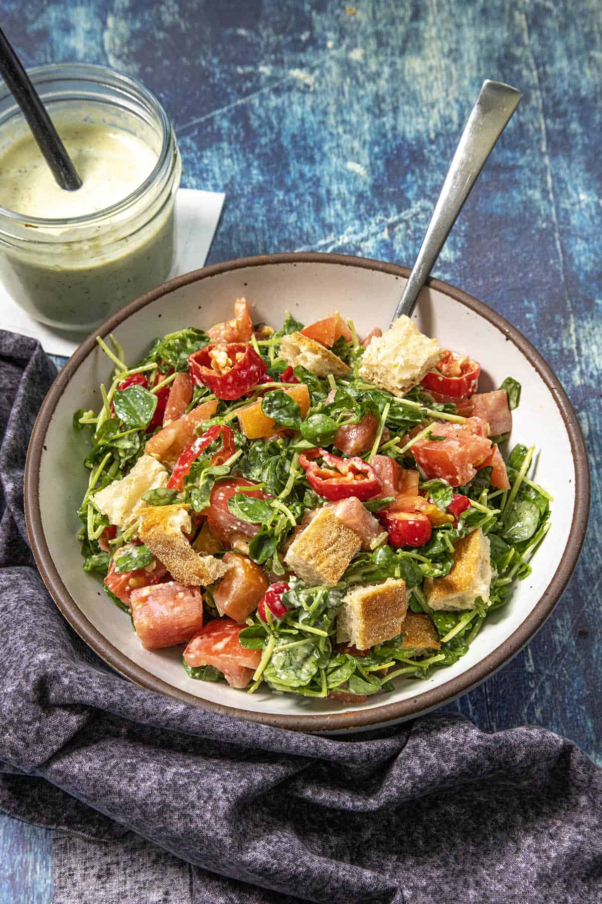 A bowl of salad with tomatoes, arugula, croutons, and dressing sits on a blue surface, next to a jar of creamy Jalapeno Ranch Dressing with a spoon and a dark cloth napkin beneath the bowl.