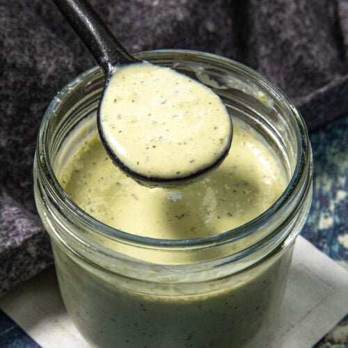 A spoon holds creamy green Jalapeno Ranch Dressing above an open glass jar, with a dark cloth and blue surface in the background.