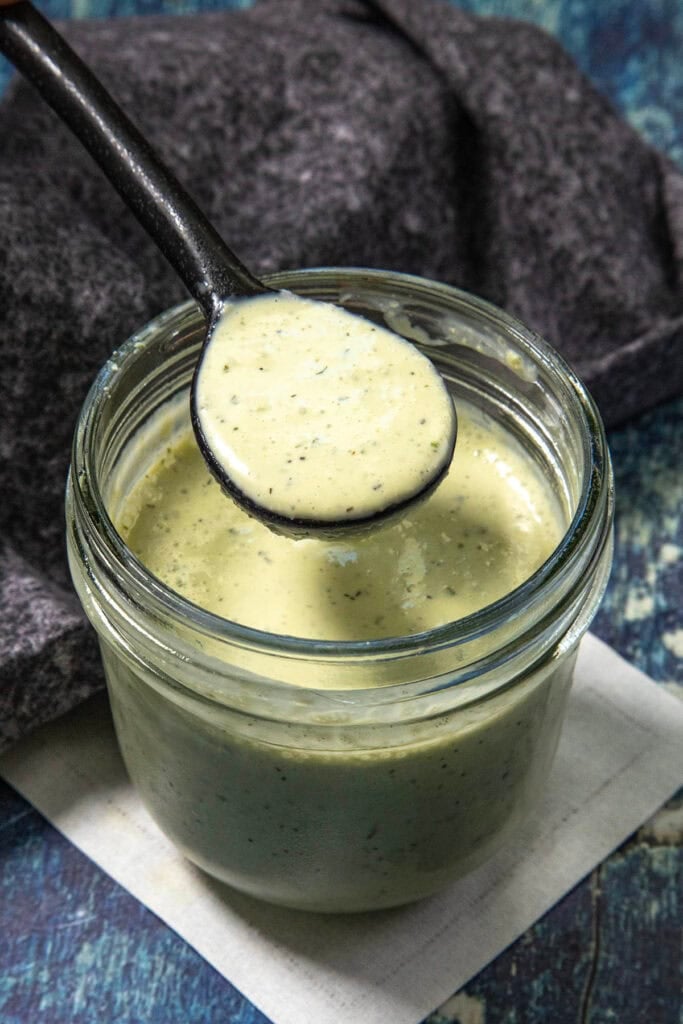 A spoon holds creamy green Jalapeno Ranch Dressing above an open glass jar, with a dark cloth and blue surface in the background.