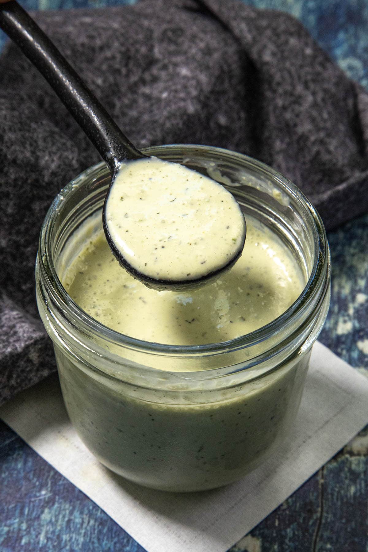 A spoon holds creamy green Jalapeno Ranch Dressing above an open glass jar, with a dark cloth and blue surface in the background.