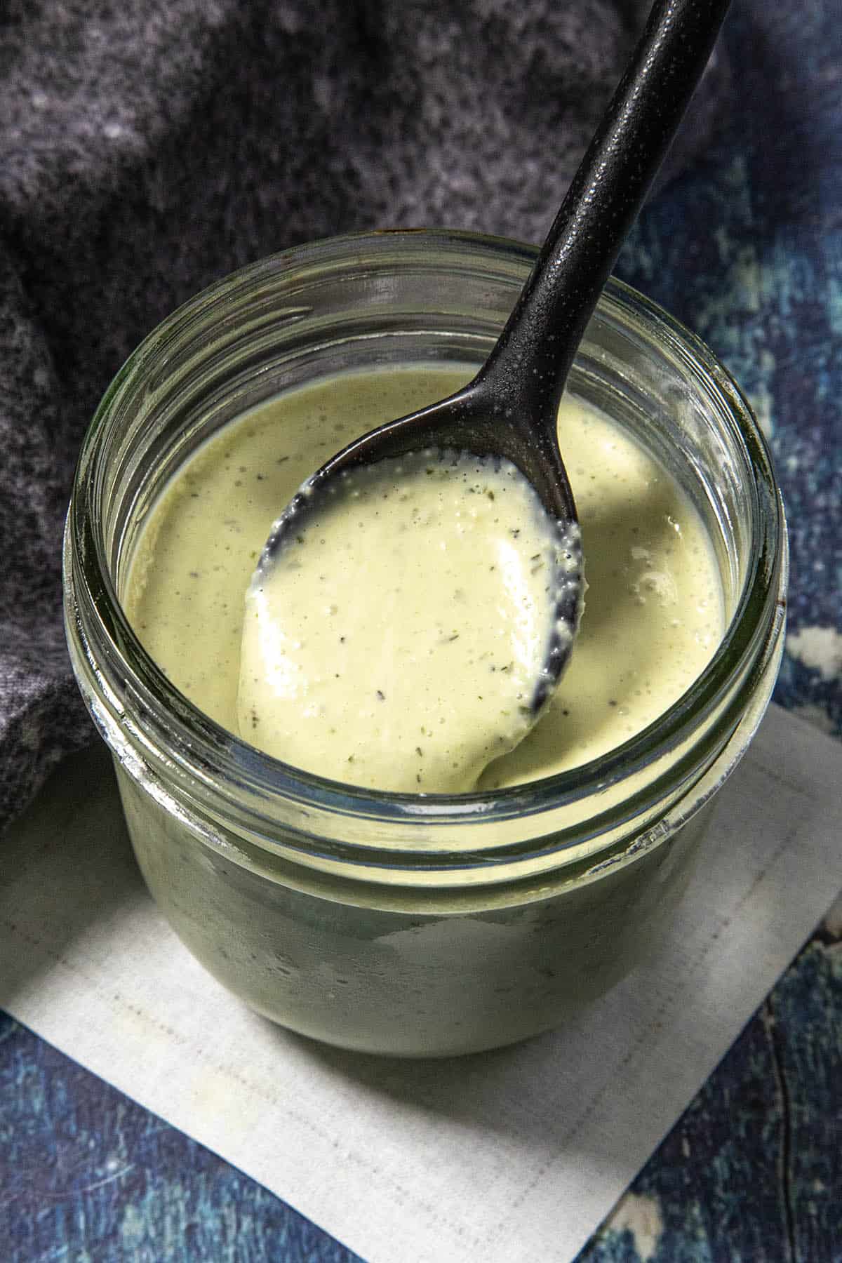 A glass jar filled with creamy green Jalapeno Ranch Dressing, with a black spoon resting on top and some sauce on the spoon. The jar sits on a white piece of paper on a blue textured surface.